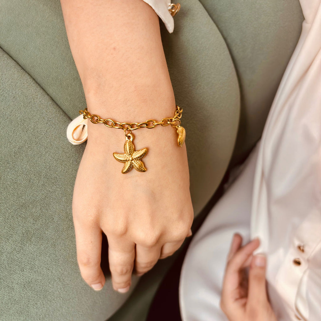 Model's hand resting on sofa wearing starfish and cowrie shell bracelet showing details on gold chain bracelet - dainty chic minimalist elegant gold plated bracelet - hypoallergenic anti‑tarnish waterproof nickel‑free lead‑free gold plated bracelet